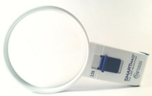 A handheld magnifying glass with a white plastic frame and a round lens. The handle is attached to the base of the lens and has some printed text on it. The text includes "SMARTMAG THE LED MAGNIFIER" and "2.5X", indicating a magnification power of 2.5 times. There is also a blue rectangular button on the handle.